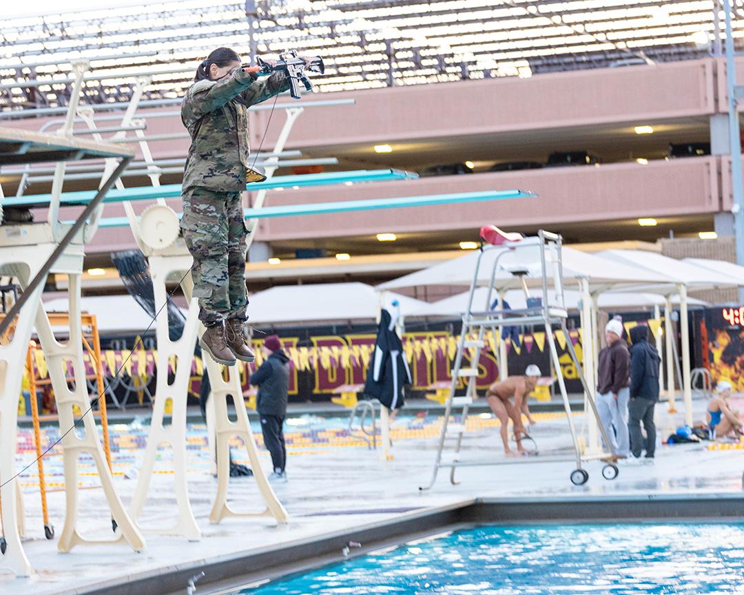An Army student wearing full camouflage and holding a fake firearm jumps into a pool. 